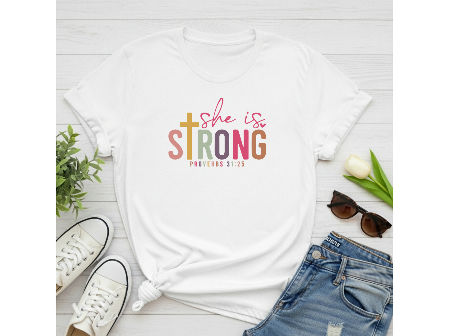White t-shirt with 'She is Strong' text on a white surface with jeans, sneakers, and sunglasses.