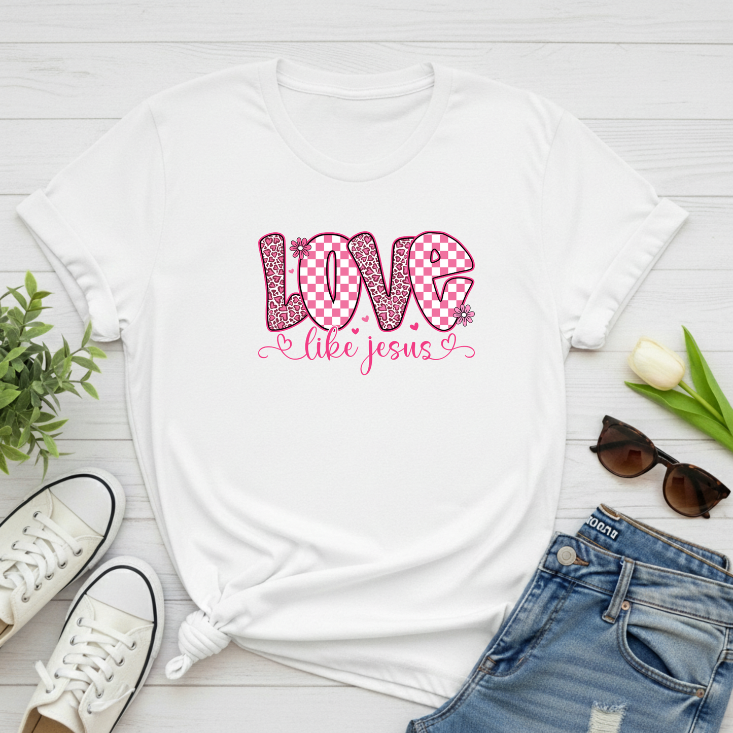 White t-shirt with 'Love like Jesus' design on a wooden surface with shoes, jeans, and sunglasses.