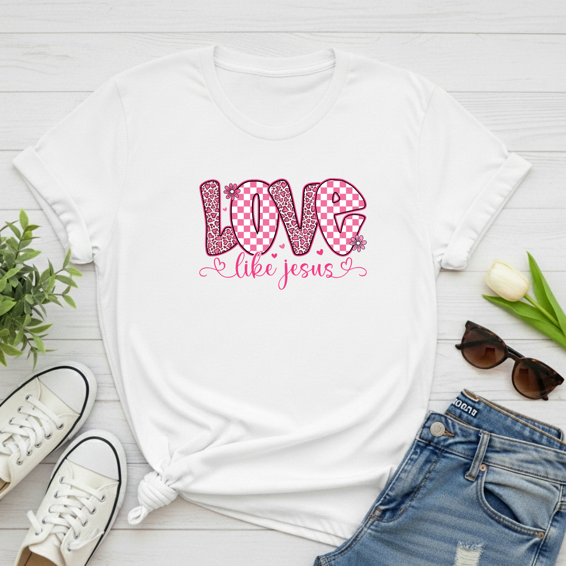 White t-shirt with 'Love like Jesus' design on a wooden surface with shoes, jeans, and sunglasses.