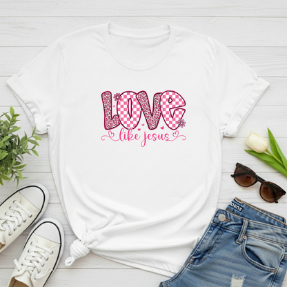 White t-shirt with 'Love like Jesus' design on a wooden surface with shoes, jeans, and sunglasses.