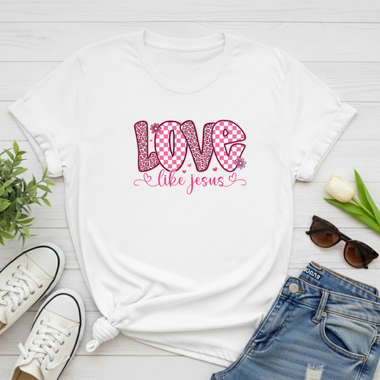 White t-shirt with 'Love like Jesus' design on a wooden surface with shoes, jeans, and sunglasses.