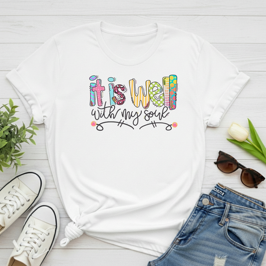 White t-shirt with colorful text 'it is well with my soul' on a white wooden surface with shoes, jeans, and sunglasses.