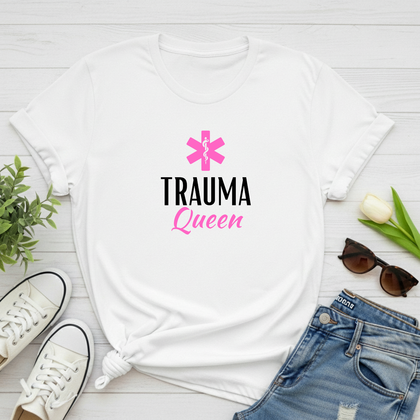 White t-shirt with 'Trauma Queen' text and pink star design on a white wooden surface with shoes, jeans, and sunglasses.