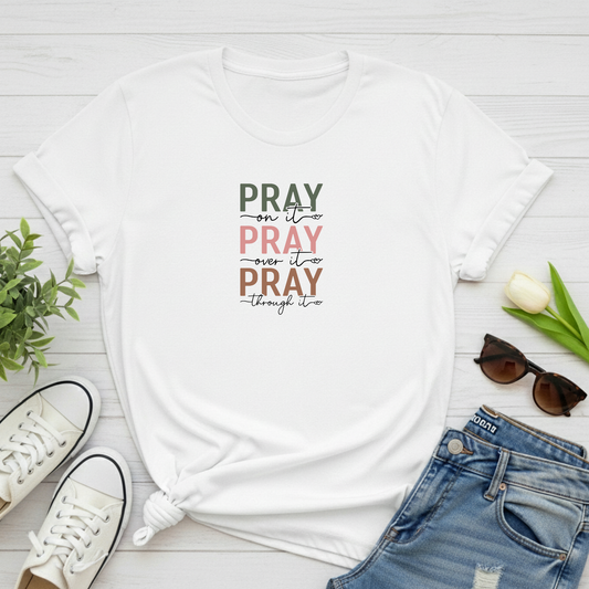 White t-shirt with 'PRAY' on it PRAY through it, PRAY over it text on a wooden surface with shoes, jeans, and sunglasses.
