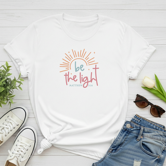 White t-shirt with 'be the light' design on a wooden surface with shoes, jeans, and sunglasses.