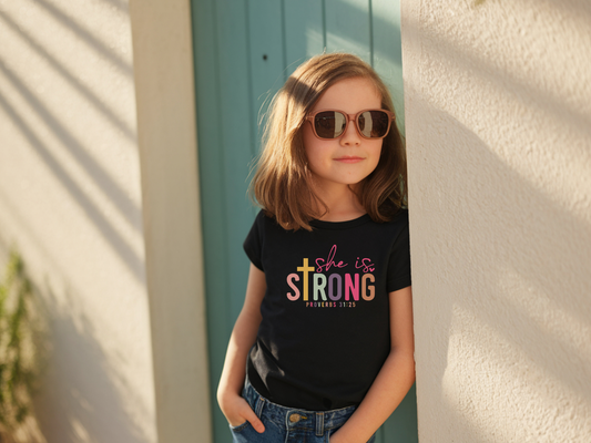 Young girl wearing sunglasses and a black t-shirt with 'She is Strong' text.