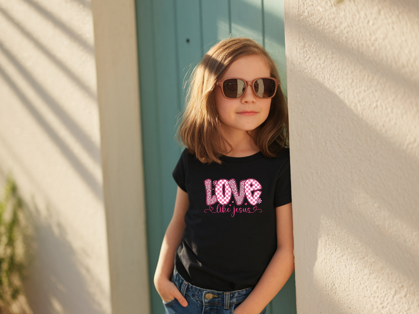 Young girl wearing sunglasses and a black t-shirt with 'LOVE LIKE JESUS' printed on it, standing against a light-colored wall.