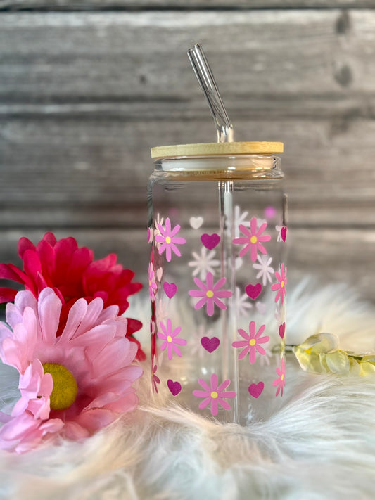 Clear tumbler with pink floral and heart pattern, gold lid, and straw on a soft surface with flowers.